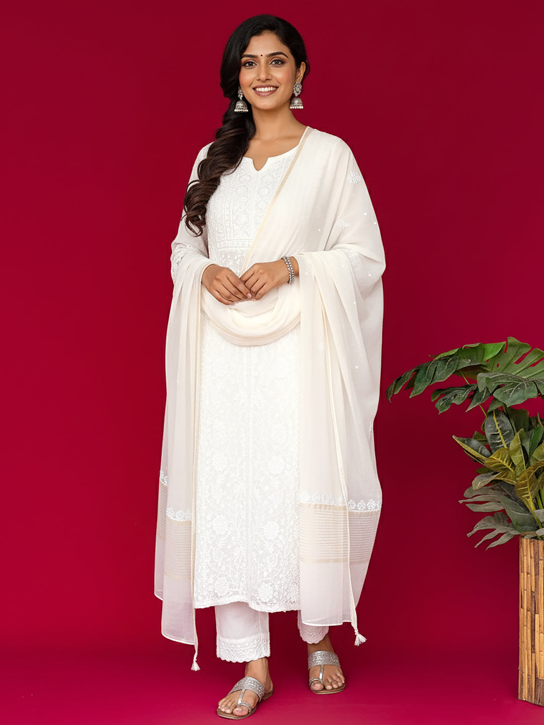 Woman in a white chikankari outfit standing against a red background with a plant on the right.